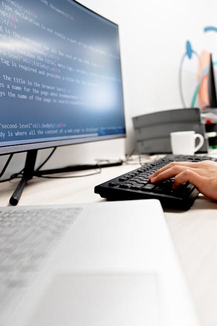 The image depicts a workspace with a person's hand operating a black keyboard in front of a large computer monitor displaying lines of code or script. To the left, part of a closed white laptop is vis