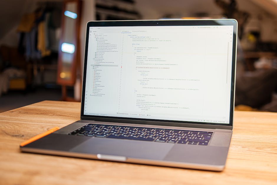 A silver laptop placed on a wooden table displaying lines of code on its screen, with a pen resting beside it. The background features a cozy, softly lit indoor space with warm-toned furniture and a m